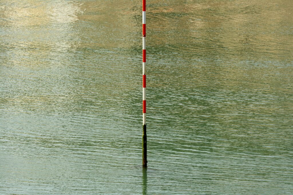 A red and white post stands in calm water, reflecting stillness and measurement.