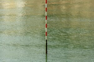 A red and white post stands in calm water, reflecting stillness and measurement.