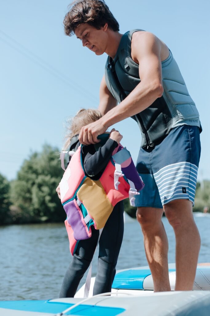 Father helps daughter prepare for wakeboarding on a sunny day by the water.